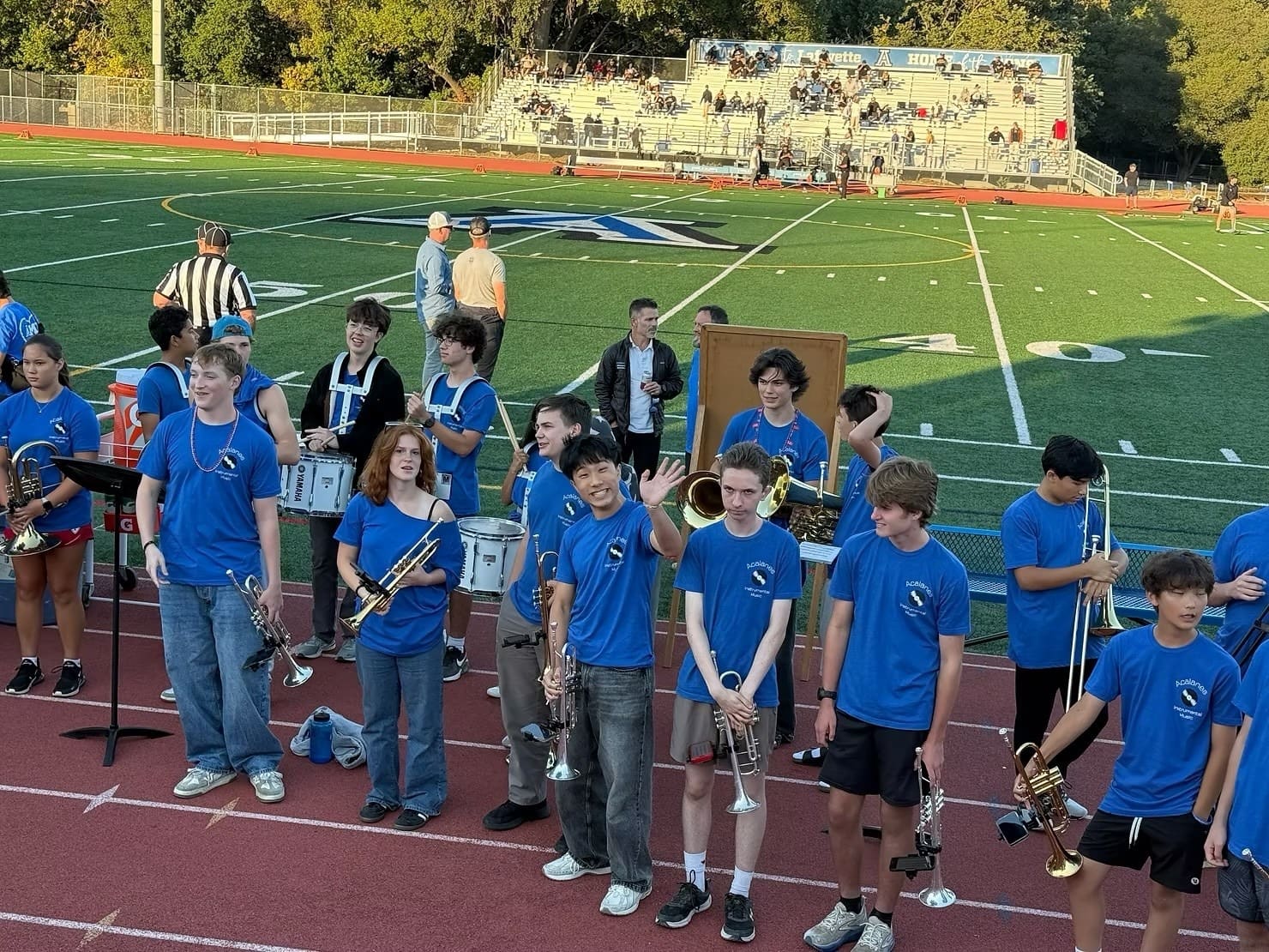 Pep Band students in blue Acalanes shirts with trumpets, trombones and drums on the track beside the football field before a game.