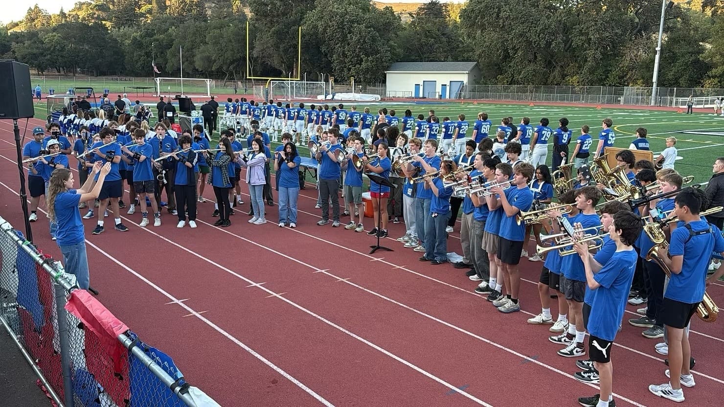 A large group of Pep Band students in blue shirts playing trumpets and other brass on the track, with the stadium behind them.
